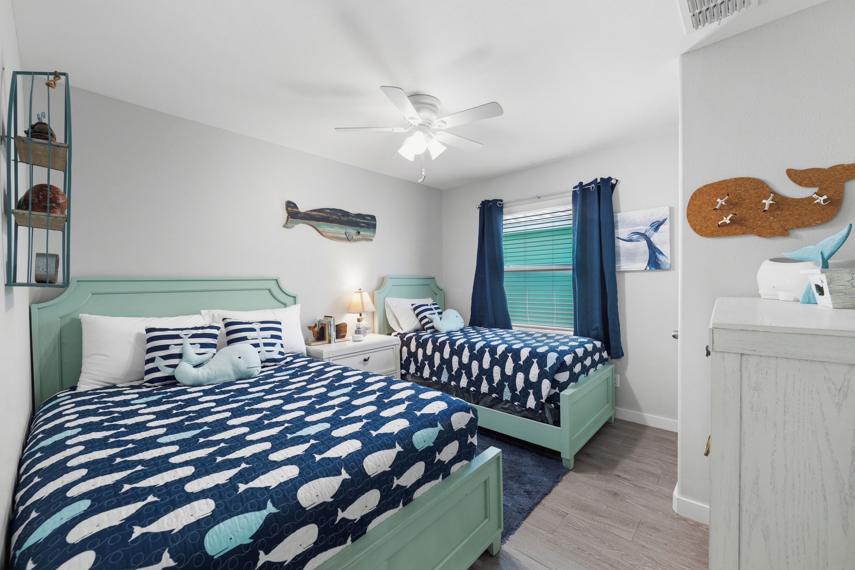 A room with two beds, a ceiling fan, a wall-mounted fish decoration, and a corkboard with bird hooks.