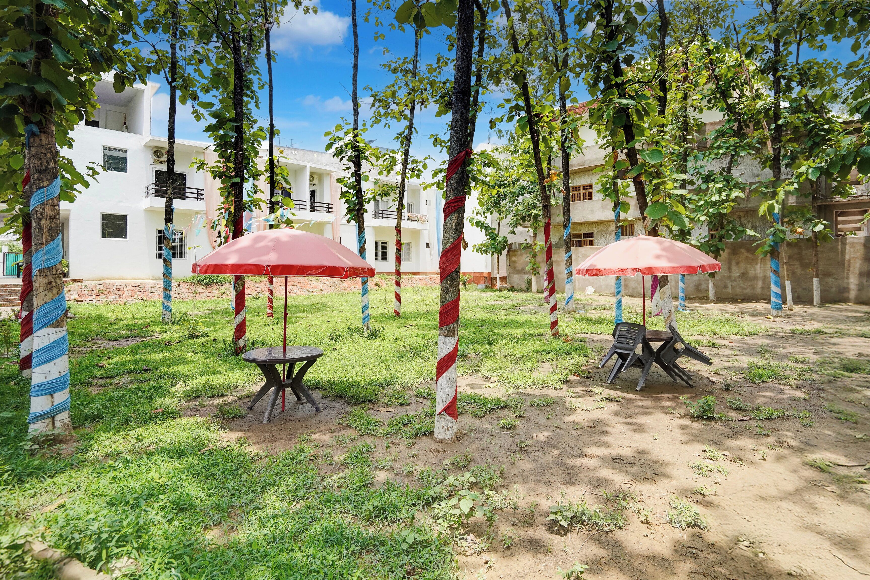 Un espacio al aire libre con dos mesas y sombrillas, rodeado de árboles y un edificio al fondo.