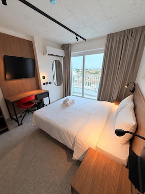 A hotel room with a bed, a desk, a TV, a mirror, and a balcony with curtains. - Campus Hub Plus  (Msida)