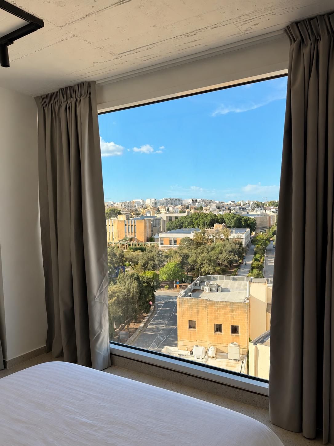 A hotel room with a large window, curtains, a bed, and a view of the cityscape.