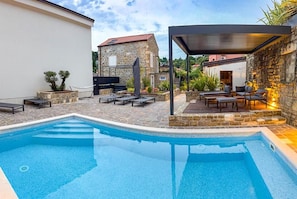 Une piscine avec une terrasse en pierre, des sièges extérieurs et un espace couvert doté d’un mur en pierre.