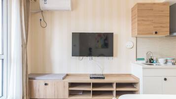 Una habitación moderna con televisor de pantalla plana en la pared, muebles de madera, un banco empotrado con espacio de almacenamiento, una pequeña cocina americana con fregadero y una ventana con cortinas.