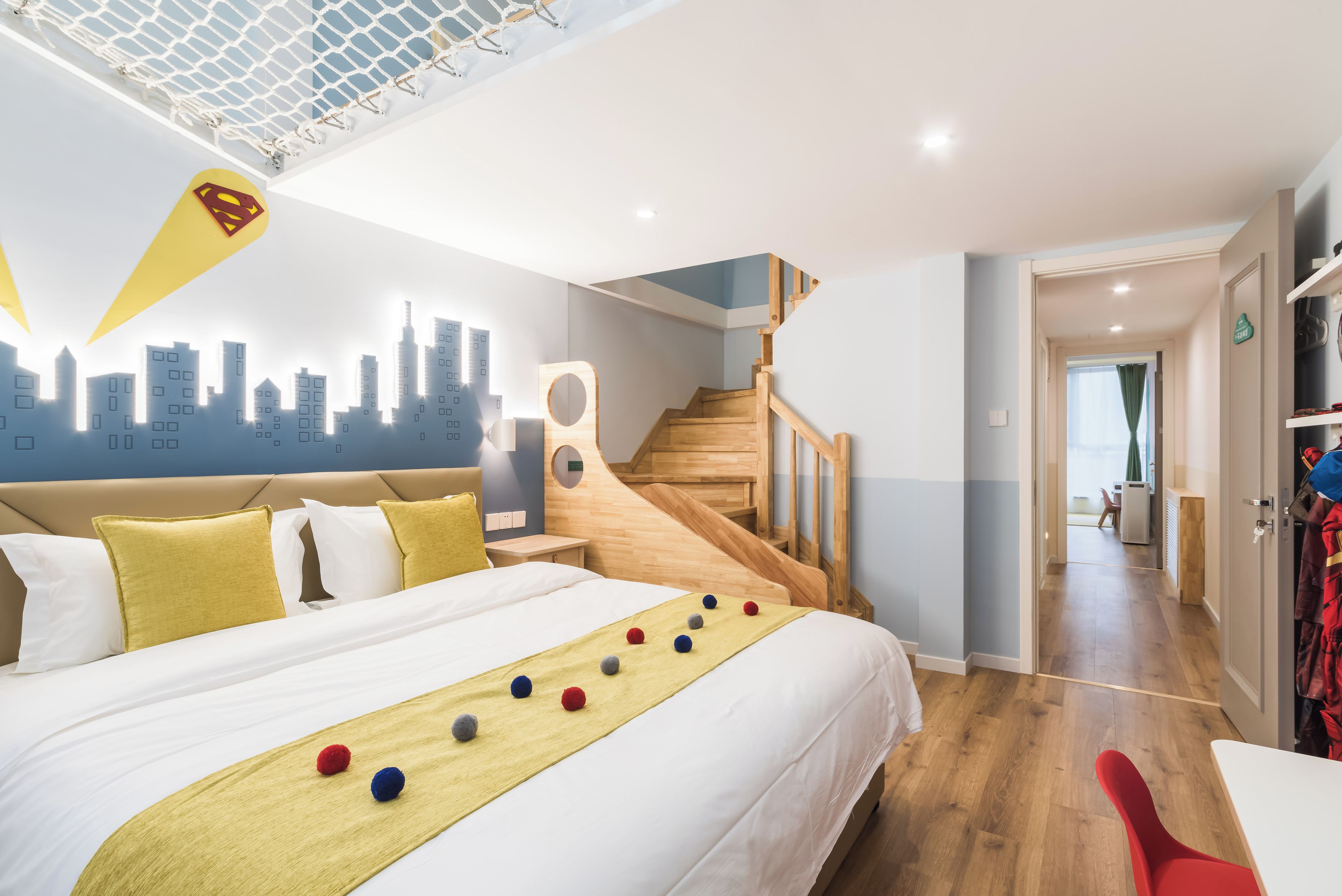 A hotel room with a bed, a cityscape mural, a wooden staircase, and a hallway leading to a dining area.