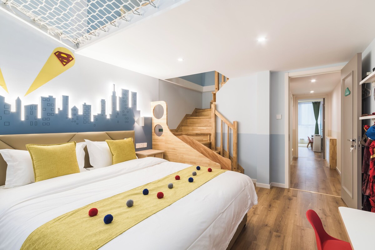 Hotellihuone, jossa on sänky, kaupunkimaisemaa esittävä seinämaalaus, puinen portaikko ja käytävä, joka johtaa ruokailutilaan.