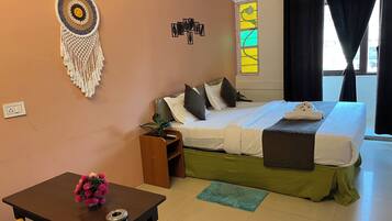 A hotel room with a bed, a bedside table with a vase of flowers, a wall-mounted dreamcatcher, a window with curtains, and a small potted plant.