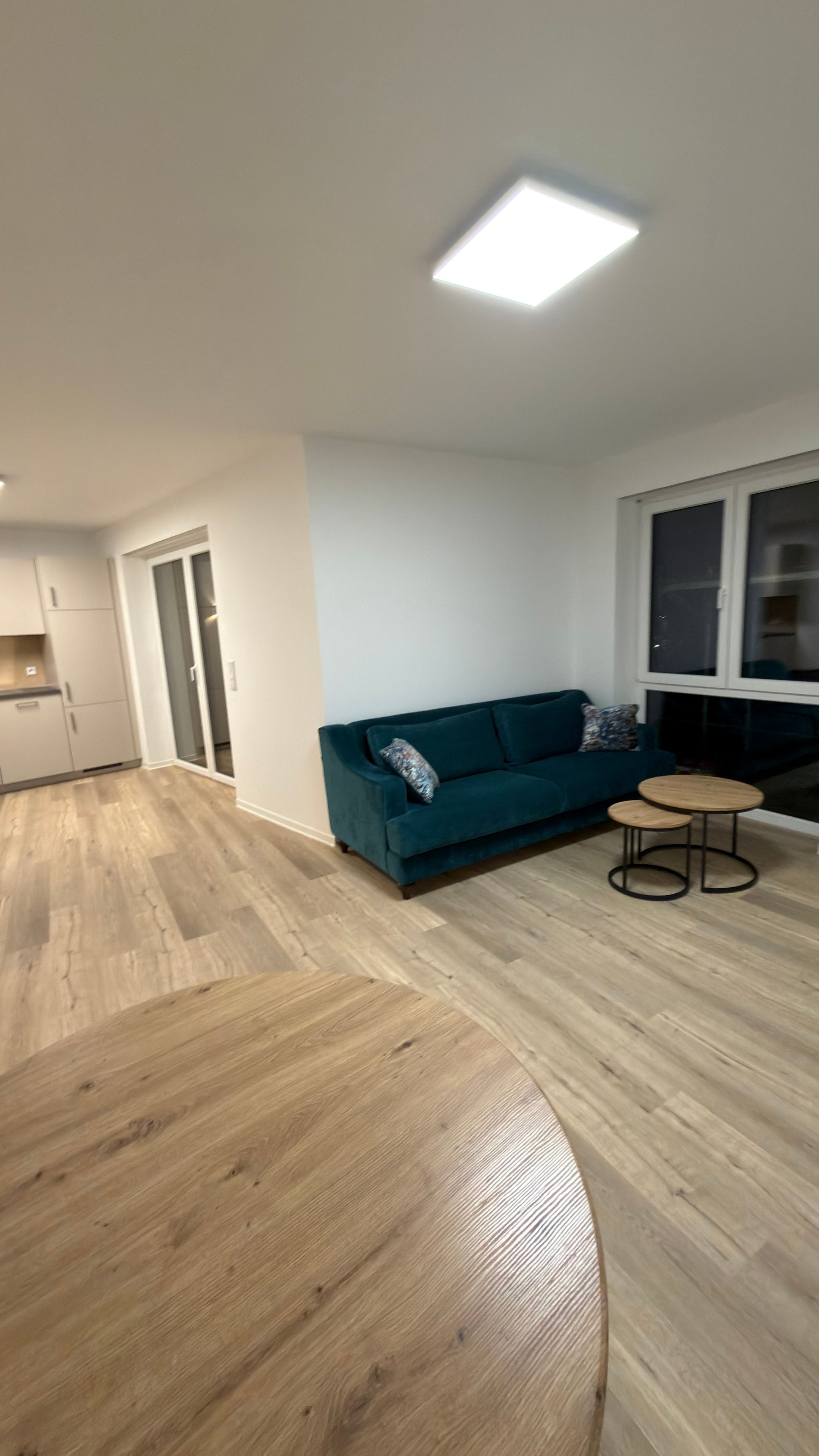 Ein modernes Wohnzimmer mit einer Couch, einem Couchtisch und einem Esstisch. Es gibt eine Küchenzeile mit Schränken und einem Kühlschrank.
