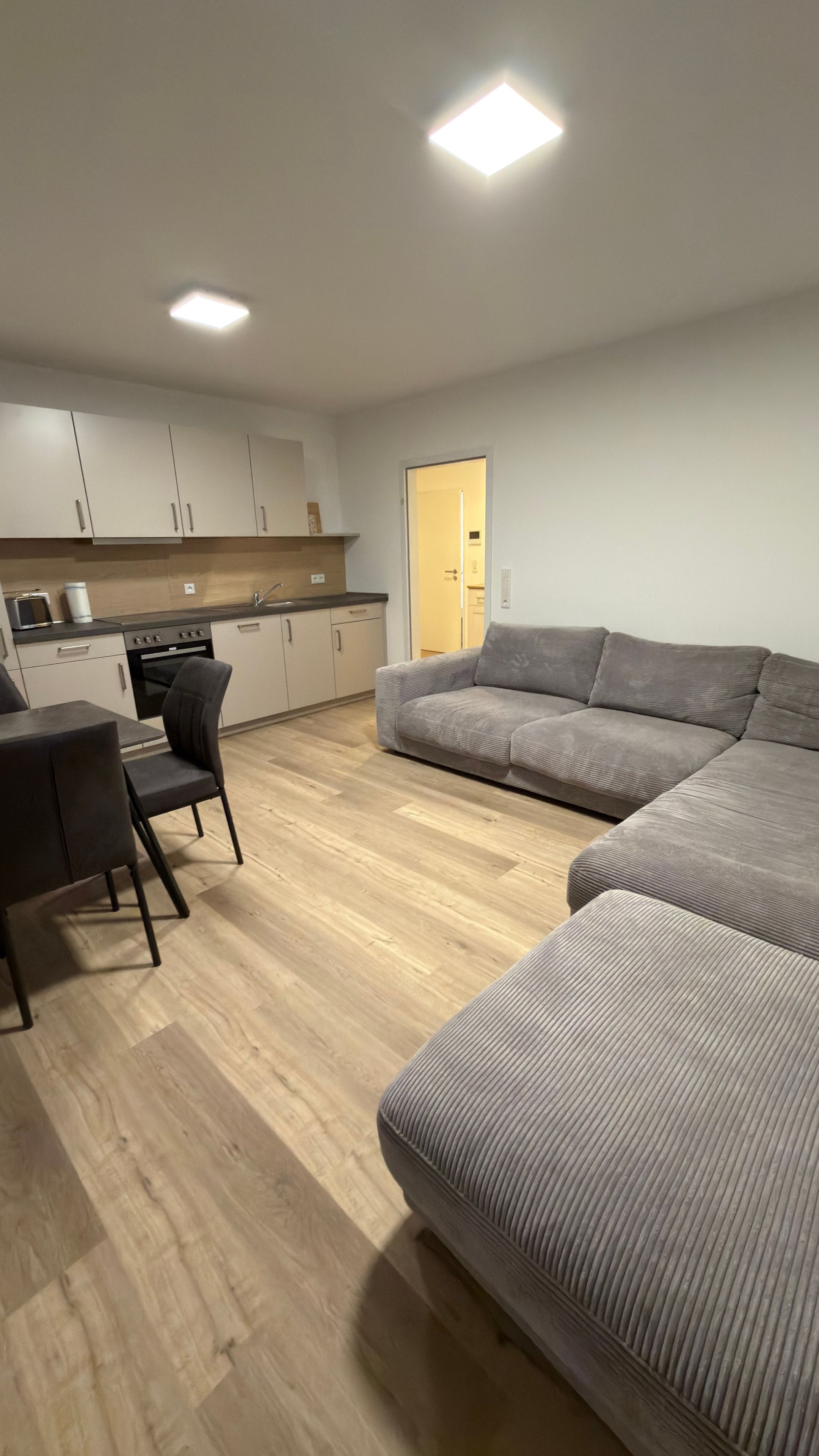 Un salon moderne avec un canapé gris, une table à manger avec des chaises et un coin cuisine équipé de meubles et d’appareils électroménagers.