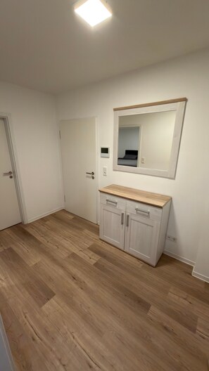 Ein Zimmer mit Holzboden, einer weißen Tür, einem weißen Schrank mit Schubladen und einem Spiegel an der Wand.