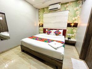 Habitación de hotel con una cama, un espejo, un equipo de aire acondicionado y una mesita de noche con lámpara.