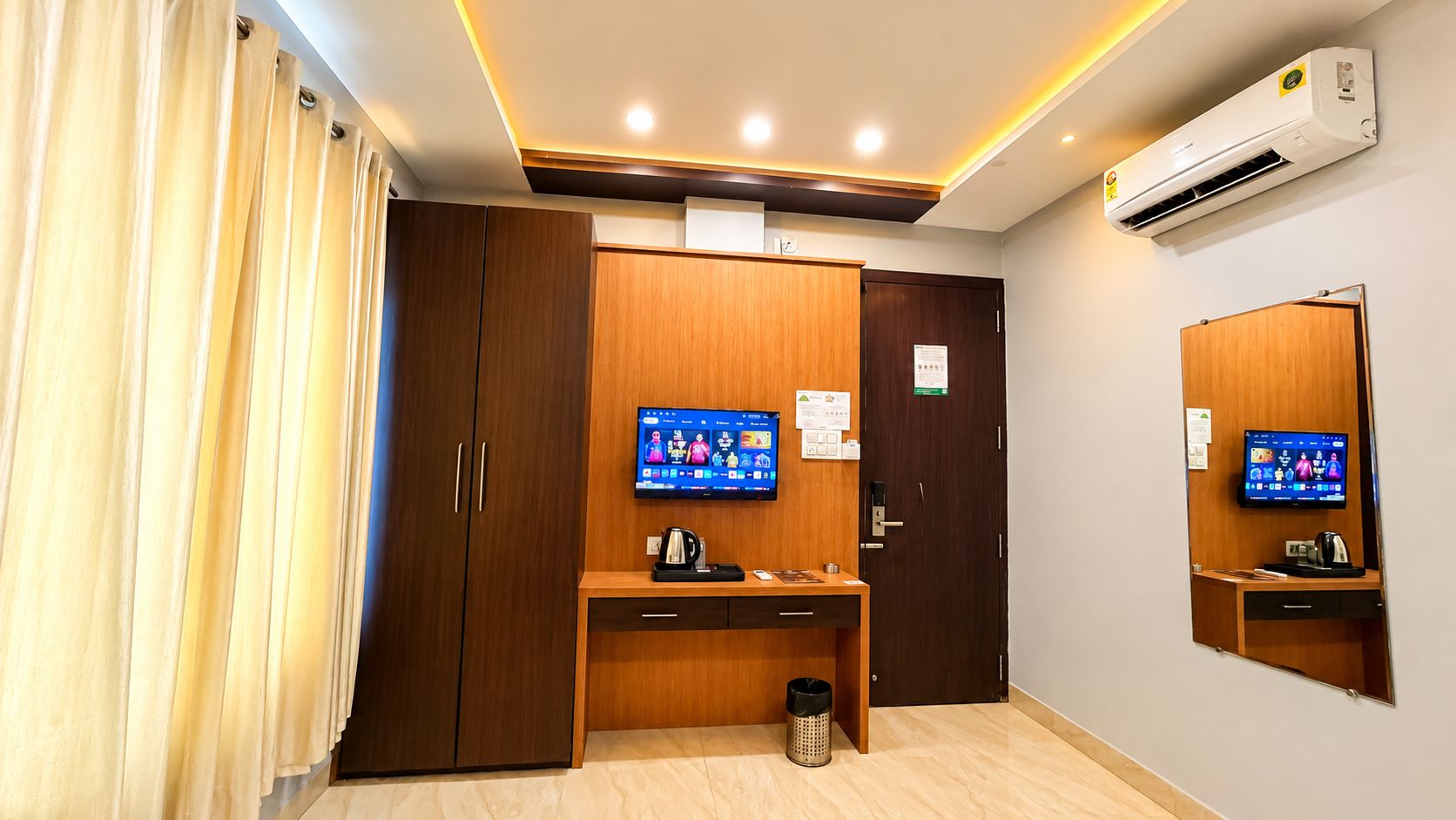 Ett hotellrum med en garderob i trä, ett skrivbord med en kaffebryggare, en papperskorg och två tv-apparater som hänger på väggen och visar information om hotellet.