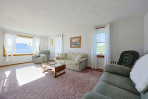 A room with a sofa, armchair, and a small table. There are two windows with curtains, and a painting on the wall.