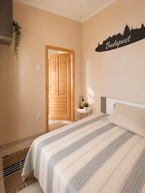 Une chambre d’hôtel avec un lit, un panneau fixé au mur sur lequel on peut lire « Budapest », une porte en bois et une petite table sur laquelle se trouve une plante.