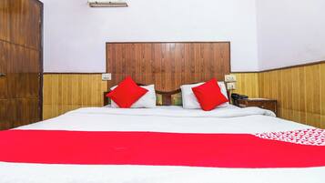 Habitación de hotel con cabecera de madera, cama con ropa blanca y almohadas rojas, mesita de noche con lámpara y equipo de aire acondicionado en la pared.