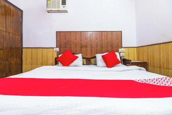 A hotel room with a wooden headboard, a bed with white bedding and red pillows, a bedside table with a lamp, and an air conditioning unit on the wall. - Hotel O Prem Chunaria (Moradabad)