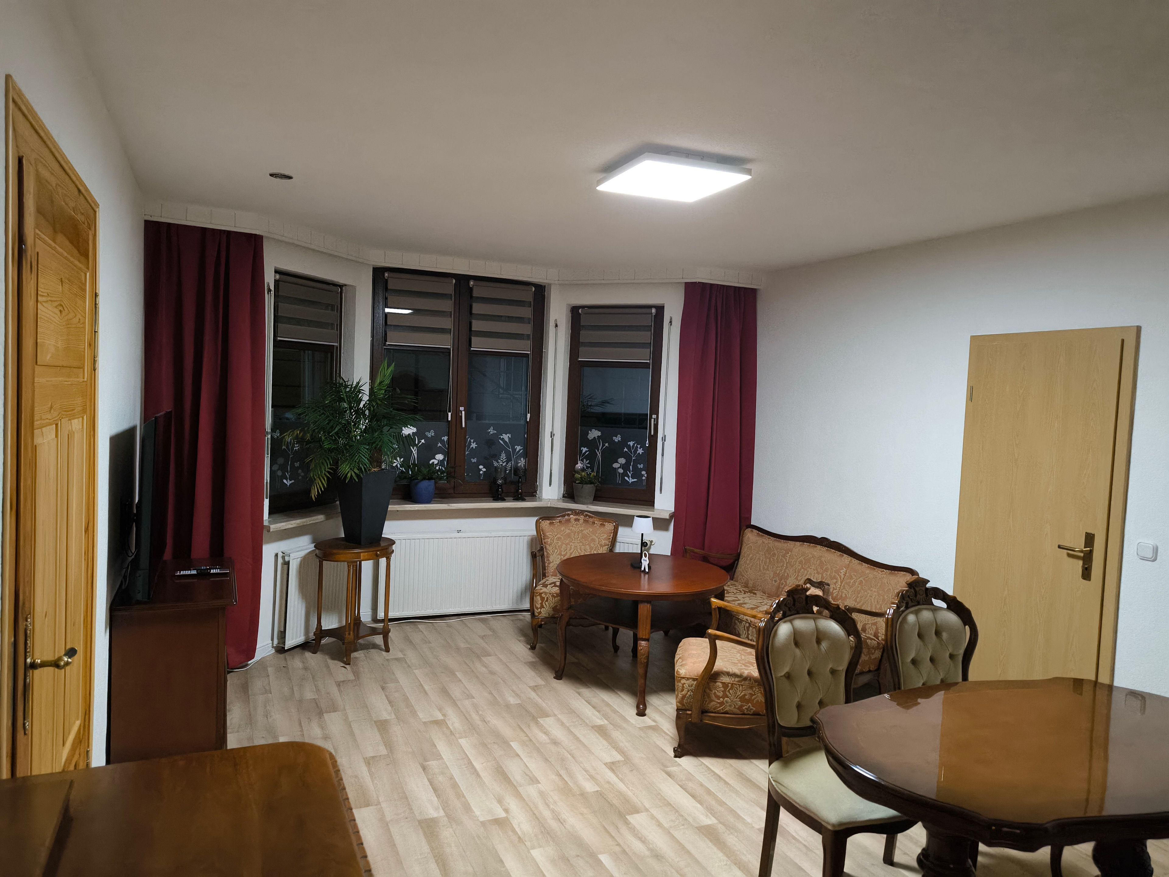 Ein Zimmer mit einem Holztisch, zwei Stühlen, einem Sofa, einer Zimmerpflanze und einem Fenster mit Vorhängen.