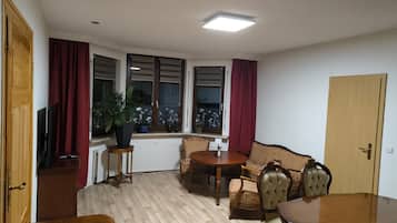 Ein Zimmer mit einem Holztisch, zwei Stühlen, einem Sofa, einer Zimmerpflanze und einem Fenster mit Vorhängen.