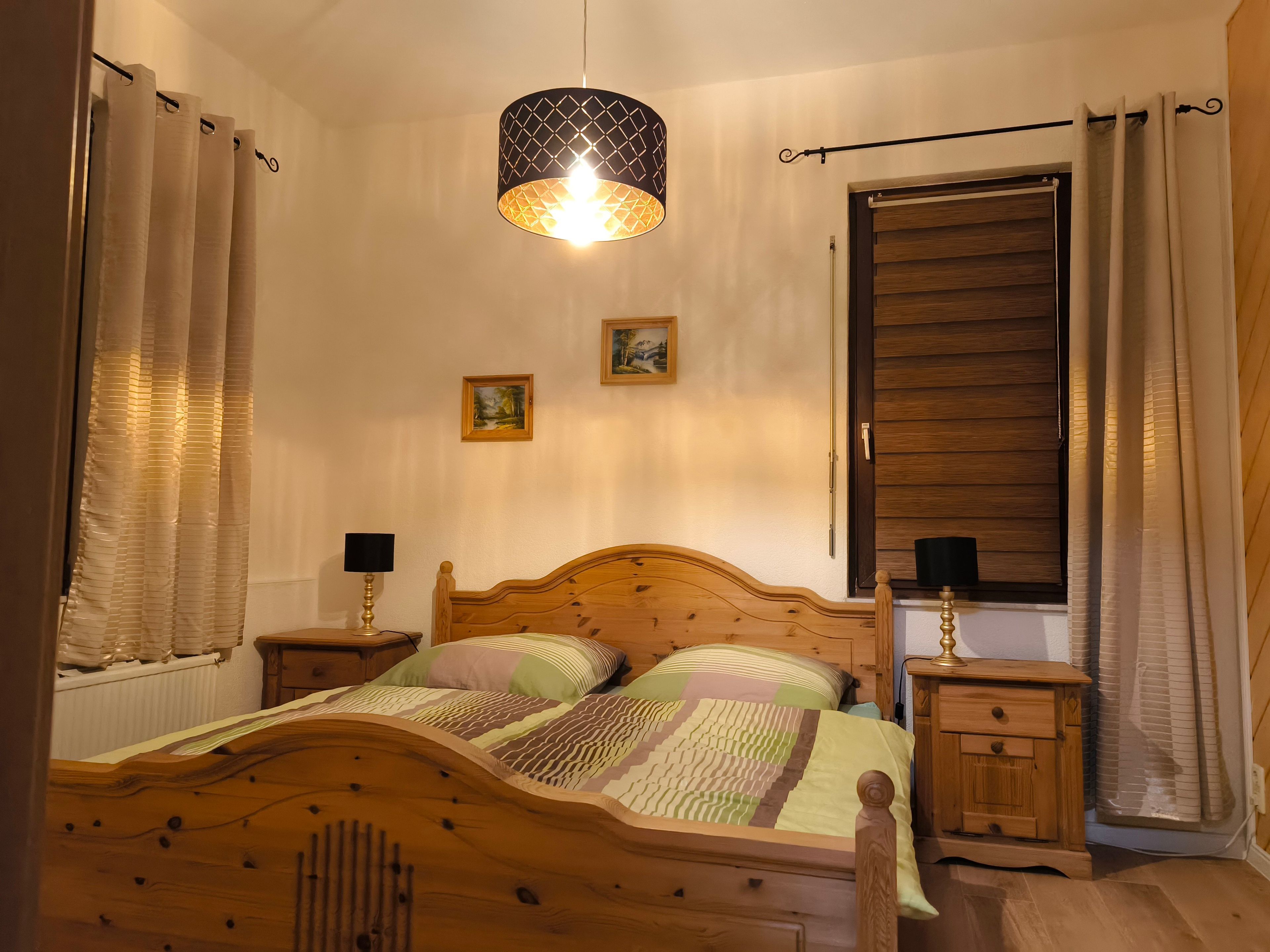 Quarto com cama de madeira, duas mesas de cabeceira com luminárias, um lustre e uma janela com venezianas de madeira.