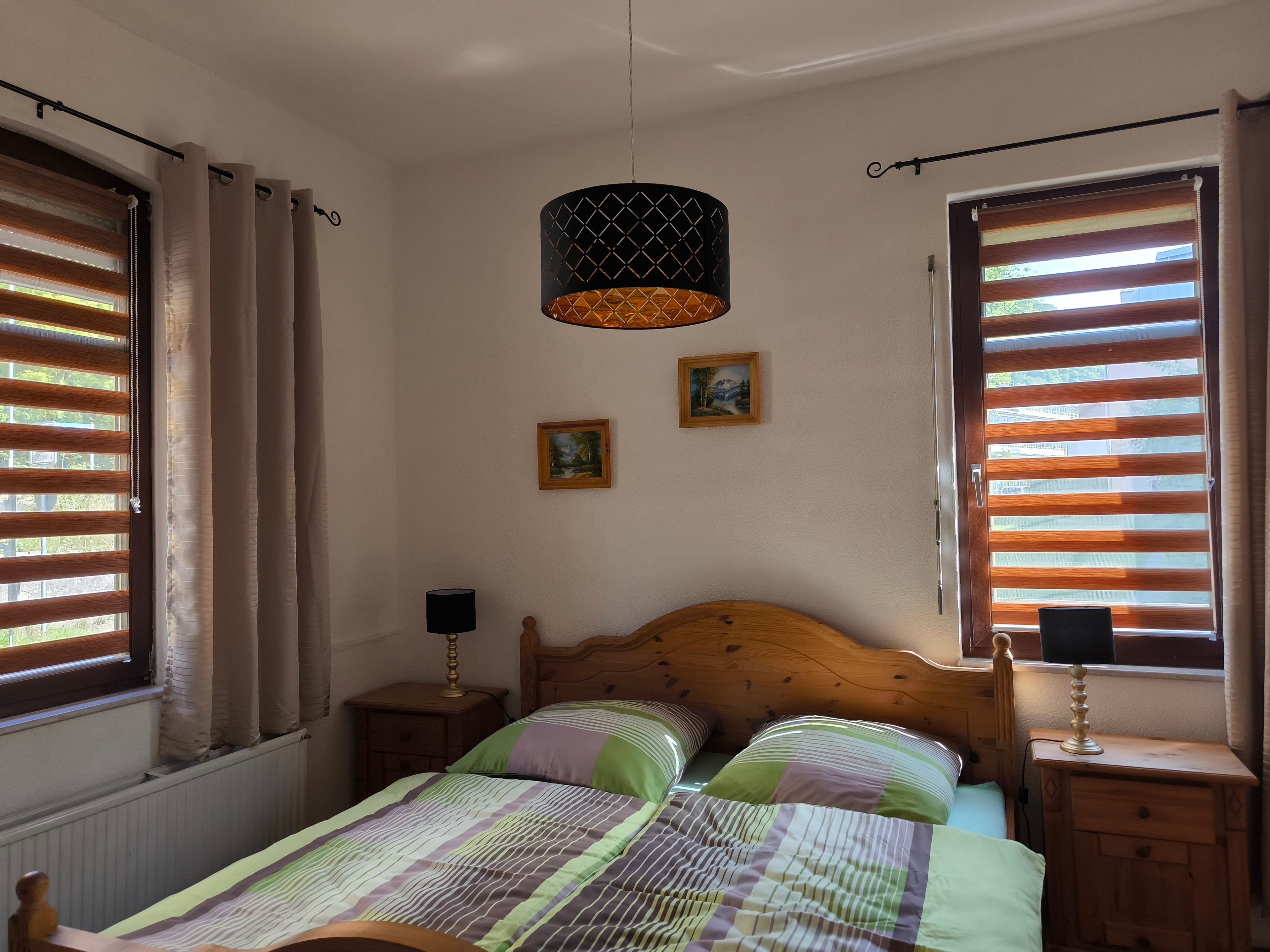 A bedroom with a wooden bed, two bedside tables, a hanging lamp, and a window with blinds.