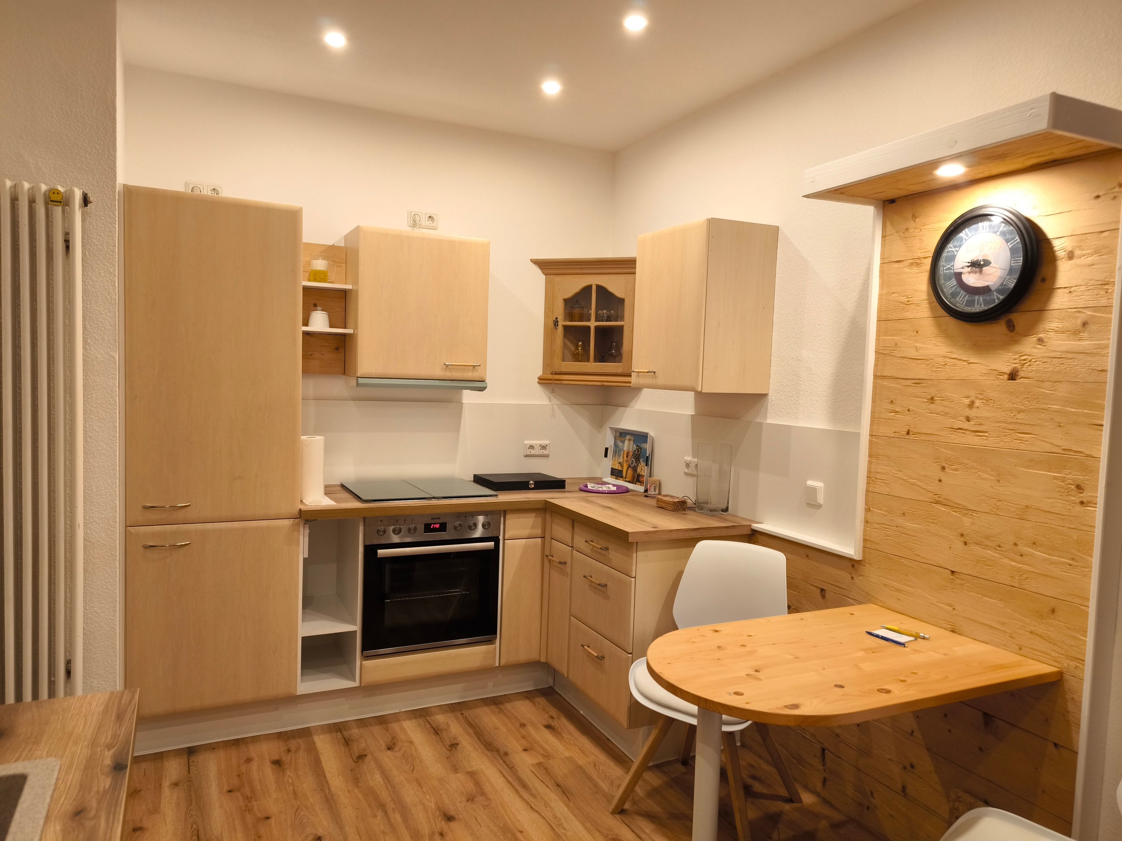 Eine kleine Küche mit Holzausstattung, einem eingebauten Backofen, einem Esstisch mit einem Stuhl und einer Wanduhr.
