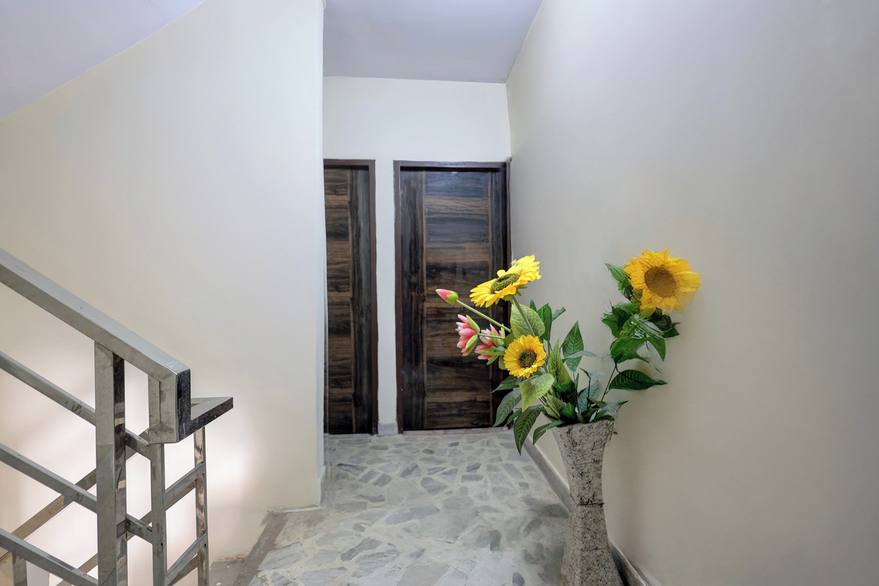 A staircase with a metal handrail, leading to a door with a dark wooden frame. A potted plant with yellow flowers is placed on the right side of the staircase.
