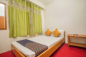 Uma cama de solteiro com colcha estampada, duas almofadas com padrão de bolinhas, mesa de cabeceira de madeira com dois controles remotos e cortinas verdes com desenho floral.