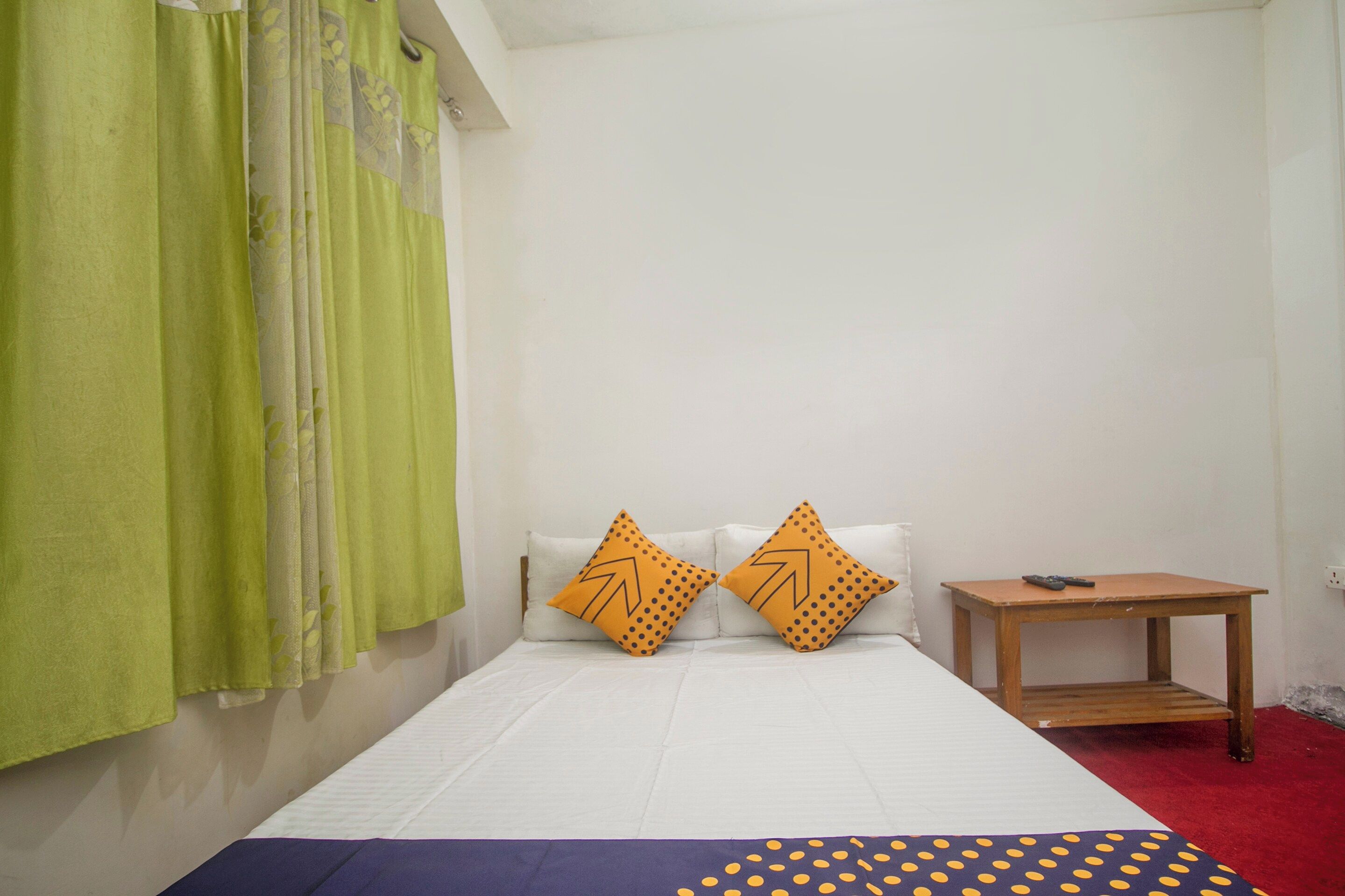 A single bed with white bedding and two orange pillows with a leaf pattern, a green curtain, and a small wooden nightstand with a remote control.
