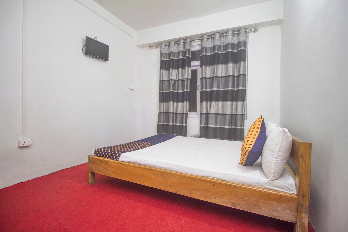 Ein Zimmer mit einem Einzelbett, einem Holzrahmen, einem roten Teppich, einem an der Wand montierten Fernseher und einem Fenster mit gestreiftem Vorhang.