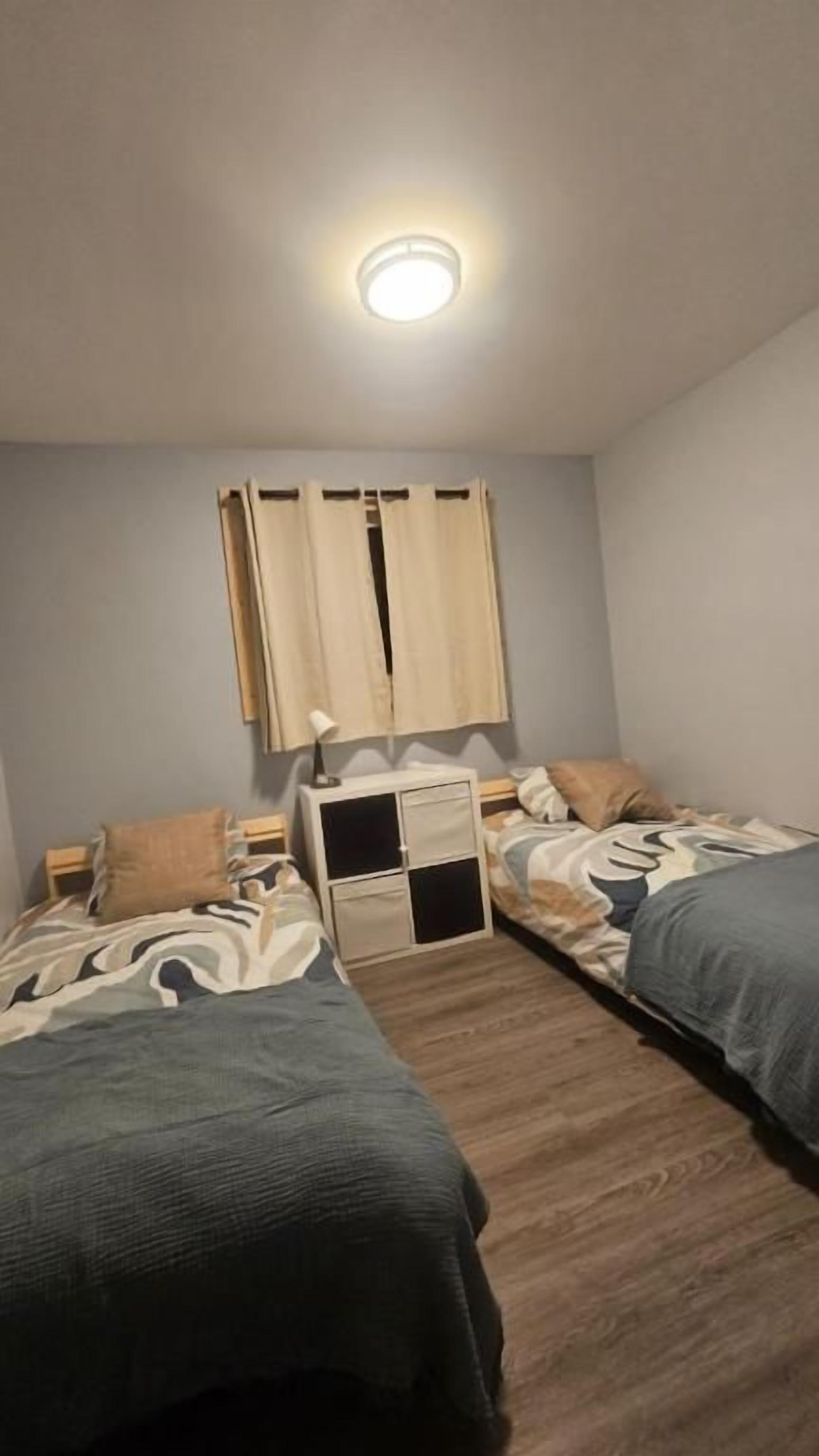 A bedroom with two beds, a bedside table with a lamp, a wardrobe, and a ceiling light.