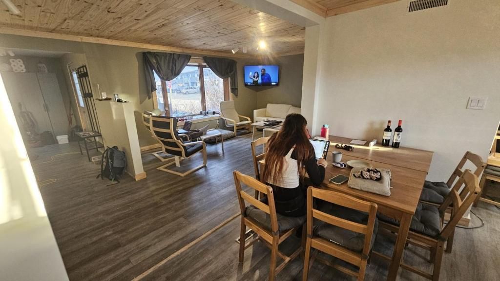 Een gezellige kamer met een houten plafond, een aan de muur bevestigde televisie, een eettafel met stoelen en een raam met gordijnen.