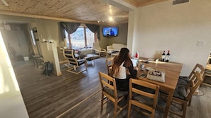 Een gezellige kamer met een houten plafond, een aan de muur bevestigde televisie, een eettafel met stoelen en een raam met gordijnen.