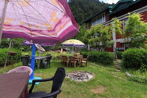 An outdoor seating area with tables and chairs, surrounded by greenery and a building with a red facade. - Hotel O Manali Meadows Home Stay (Manali)