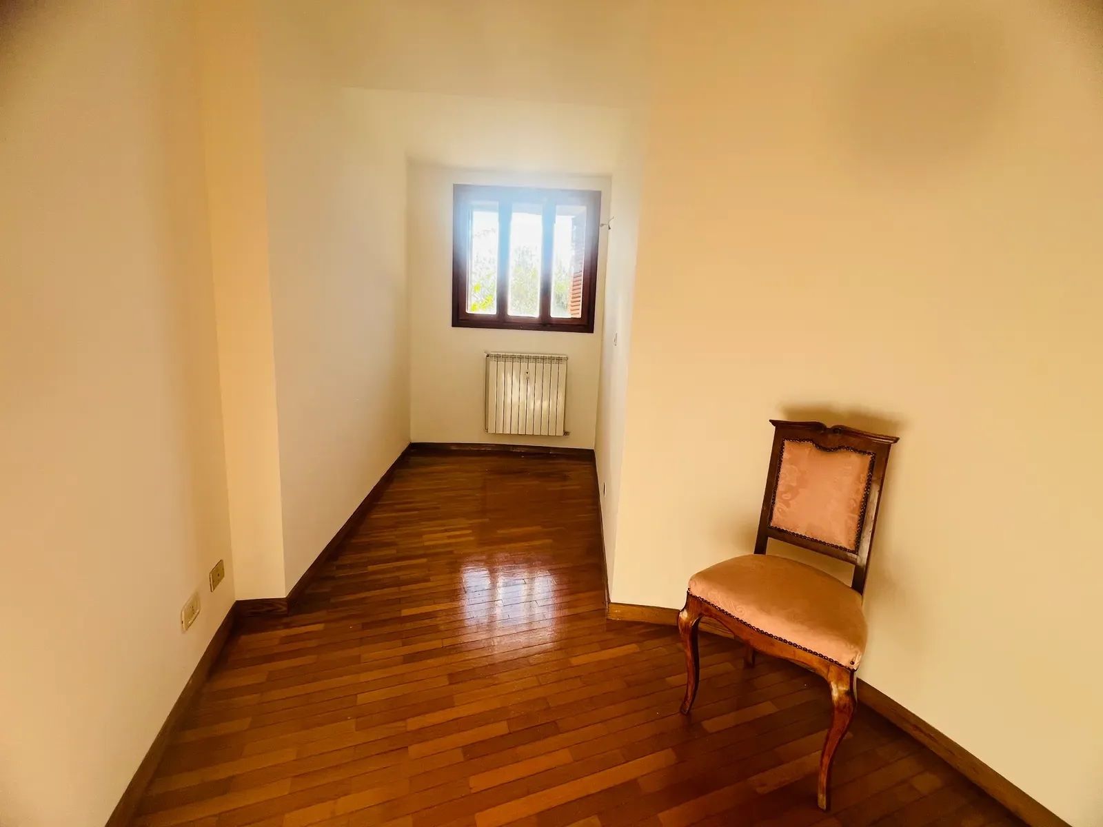 A room with wooden flooring, a radiator, a window with blinds, and a wooden chair.