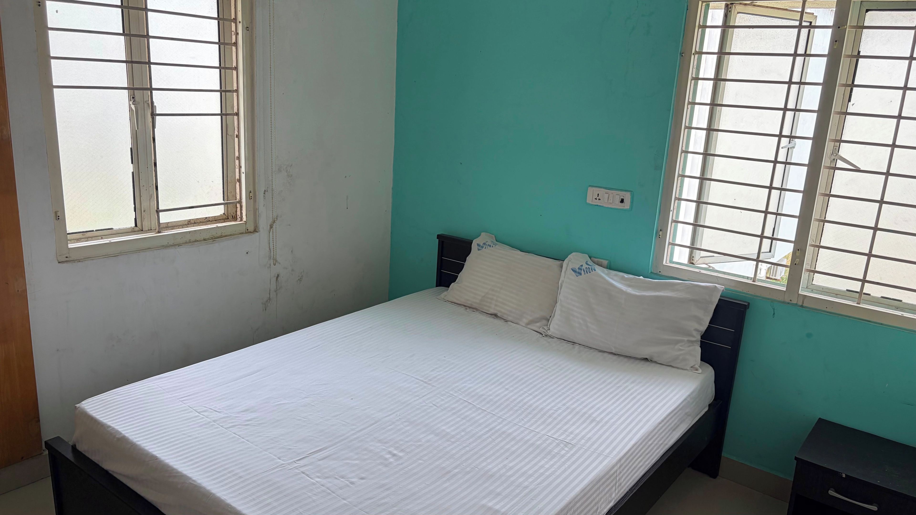 A single bed with a white mattress and pillows, a nightstand, and a window with metal bars.