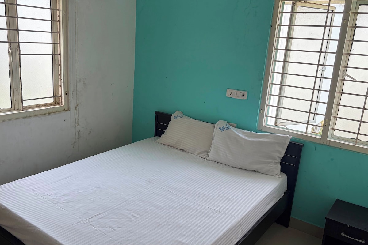 A single bed with a white mattress and pillows, a nightstand, and a window with metal bars.
