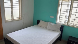 A single bed with a white mattress and pillows, a nightstand, and a window with metal bars.