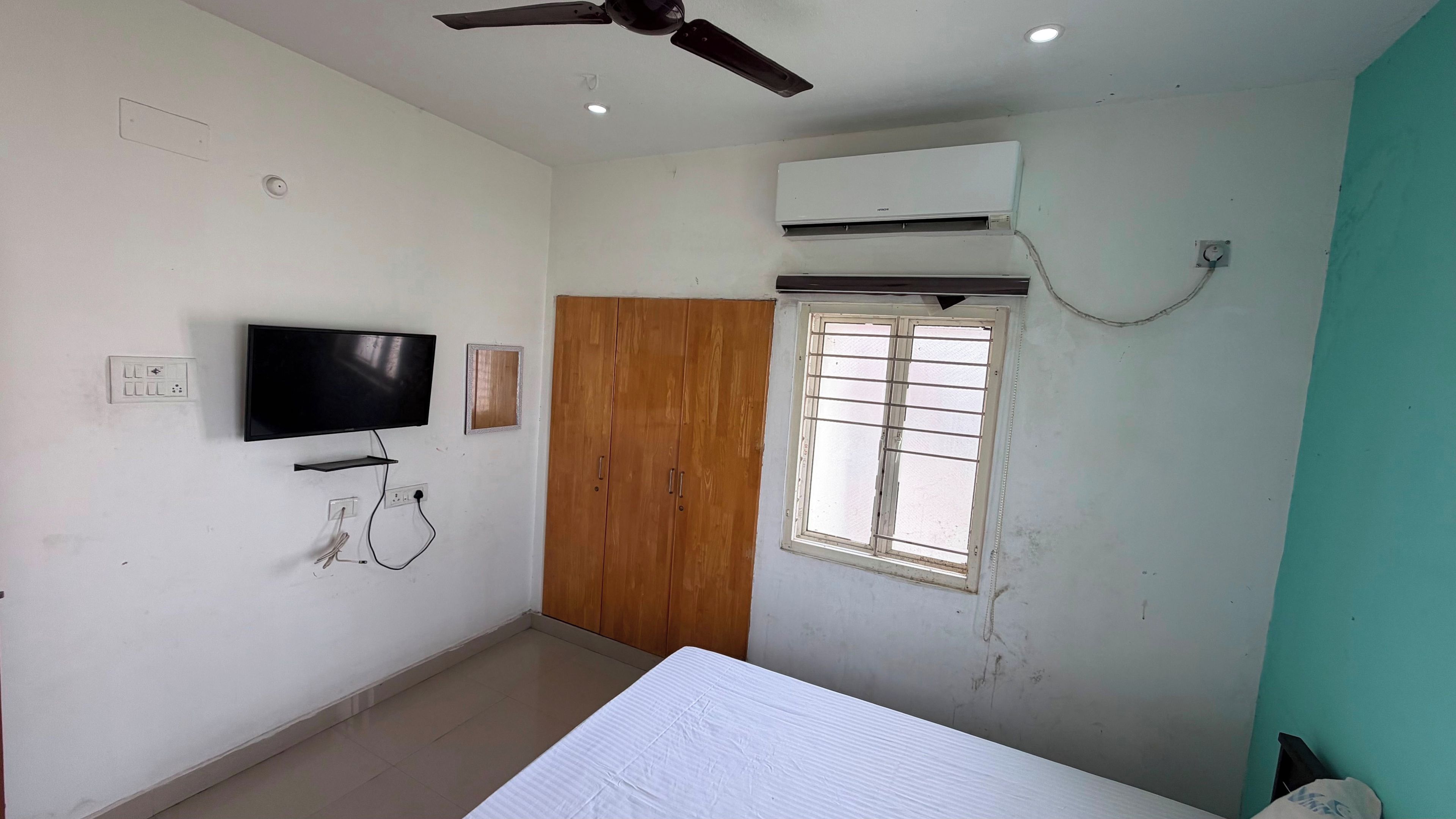 A room with a ceiling fan, an air conditioning unit, a television mounted on the wall, a wooden wardrobe, and a window with bars.