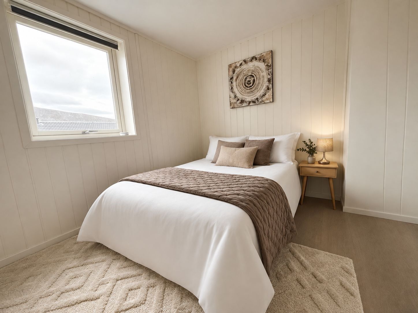 A bedroom with a bed, a bedside table, a lamp, a window, and a wall art piece.