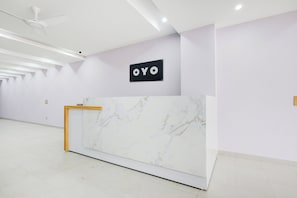 Un mostrador de recepción con encimera de mármol, un ventilador de techo y un cartel con las letras 'OYO' en una pared color lila claro.