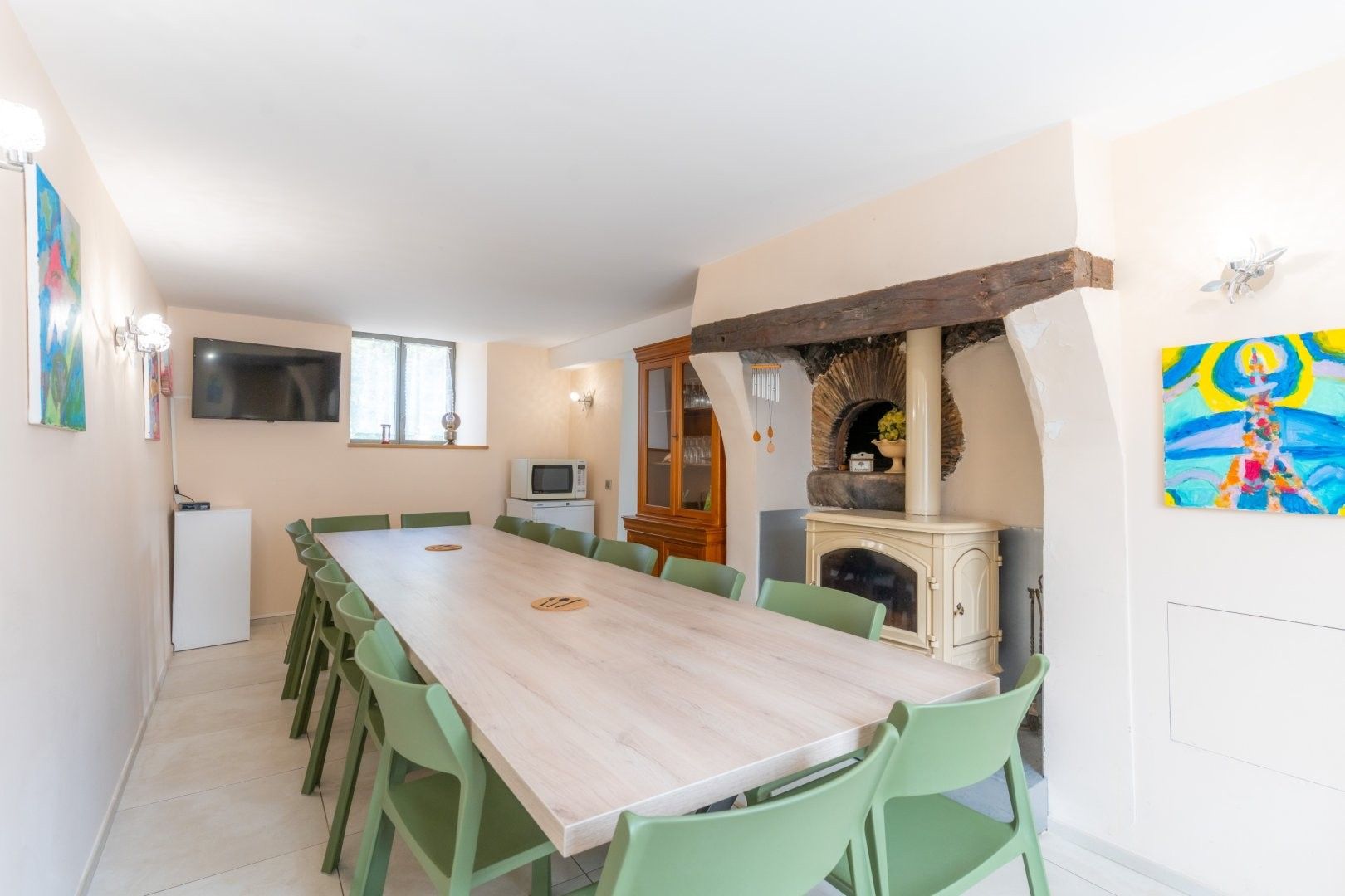 A dining area with a long table, green chairs, a television, a microwave, and a fireplace.