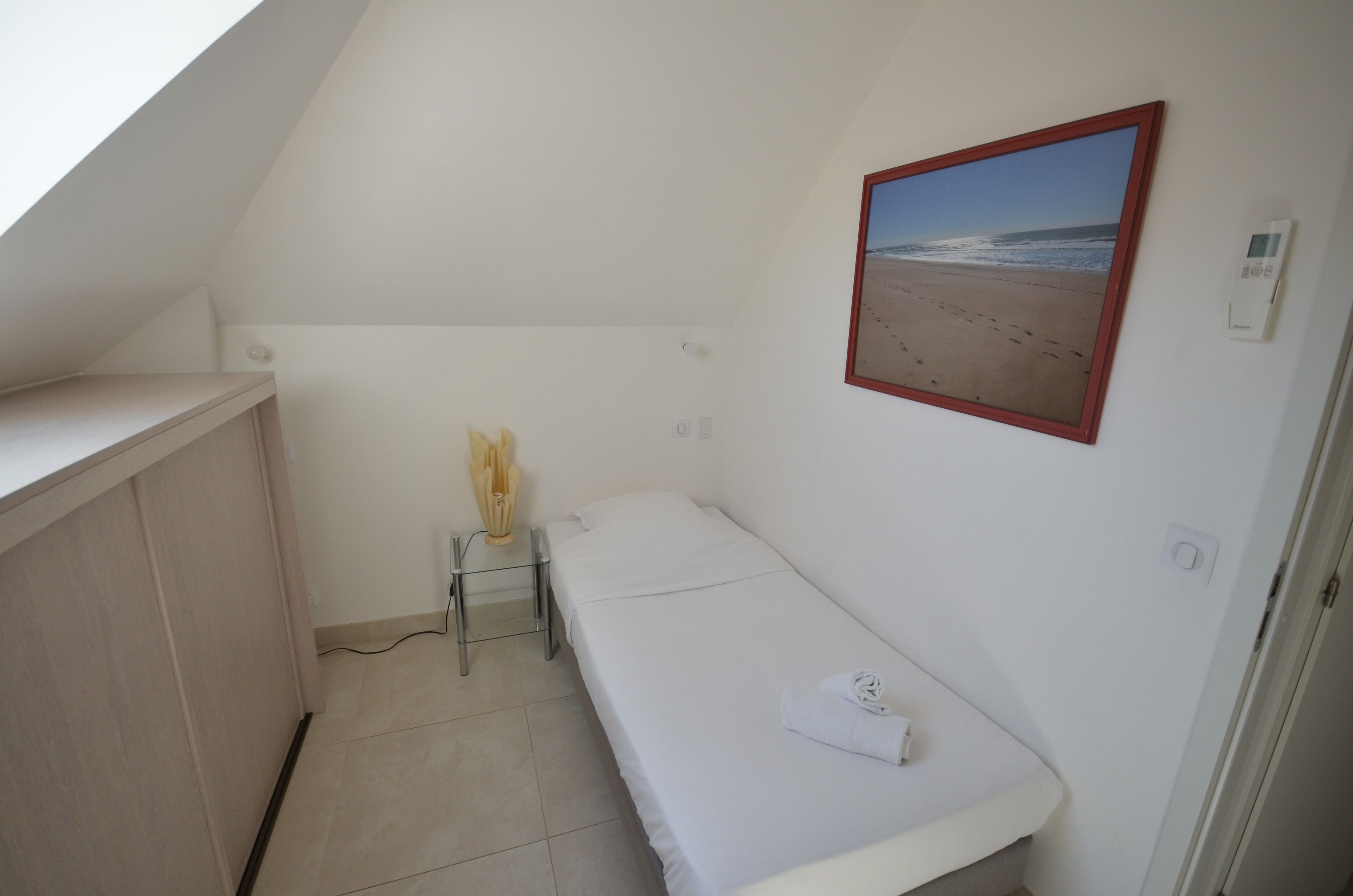 Een kleine kamer met een enkel bed en schuine dak, een nachtkastje met lamp, een ingelijst strandbeeld en een thermostaat aan de muur.