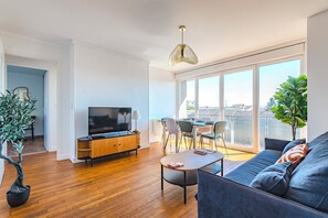Un salon moderne doté d’un téléviseur à écran plat, de parquet, d’un canapé, d’une table à manger avec des chaises et de grandes fenêtres offrant une vue sur la ville.