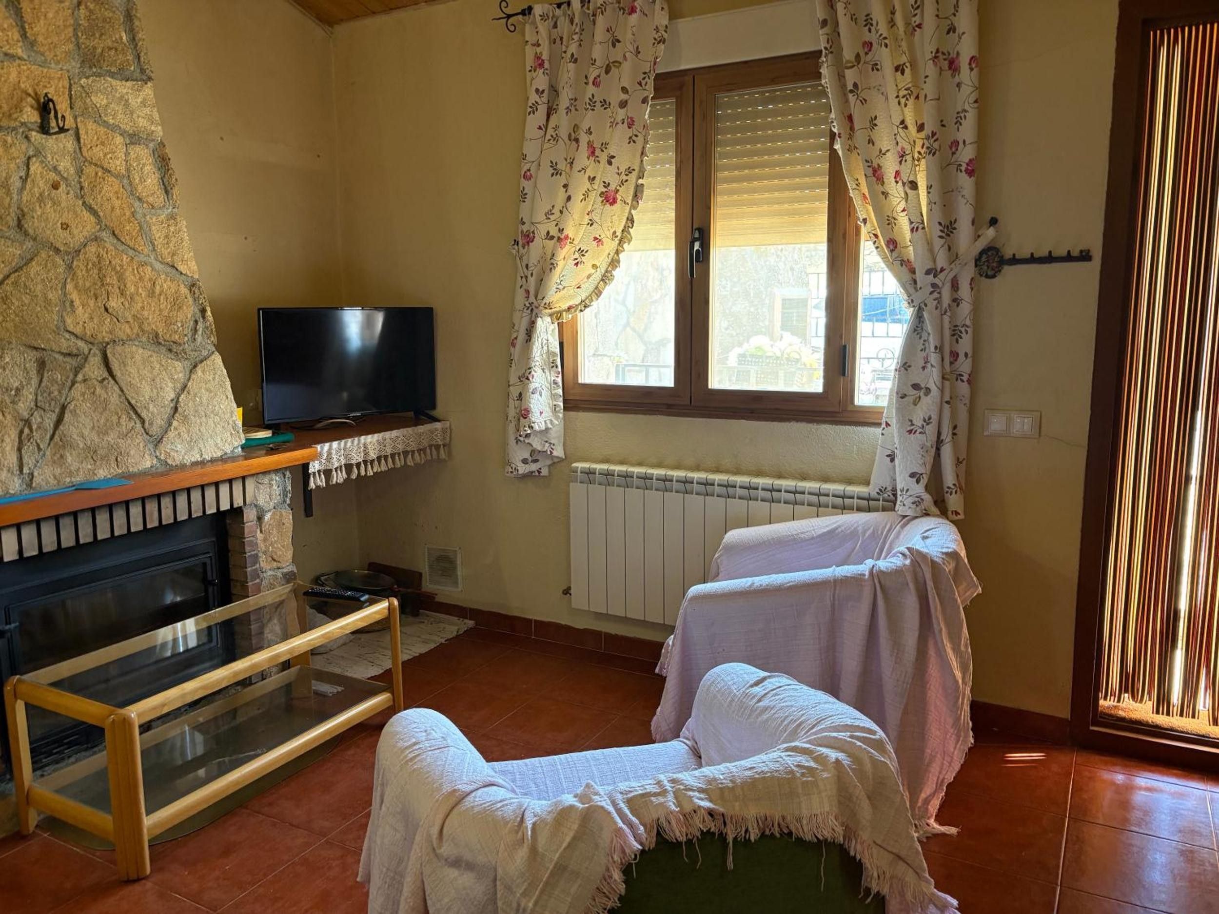 A cozy room with a stone wall, a fireplace, a television, two beds with floral curtains, and a window with blinds.