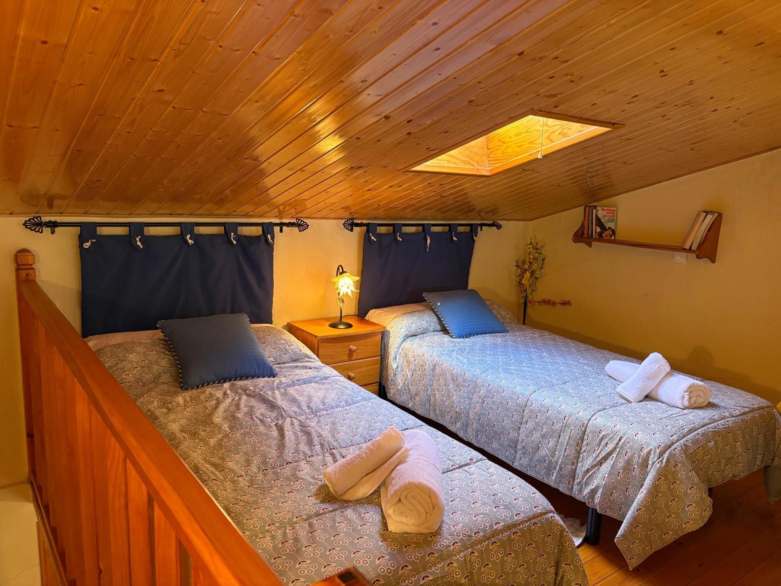 Ein Zimmer mit zwei Betten, einem Nachttisch, einer Lampe, einem Bücherregal und einem Fenster mit Vorhängen.