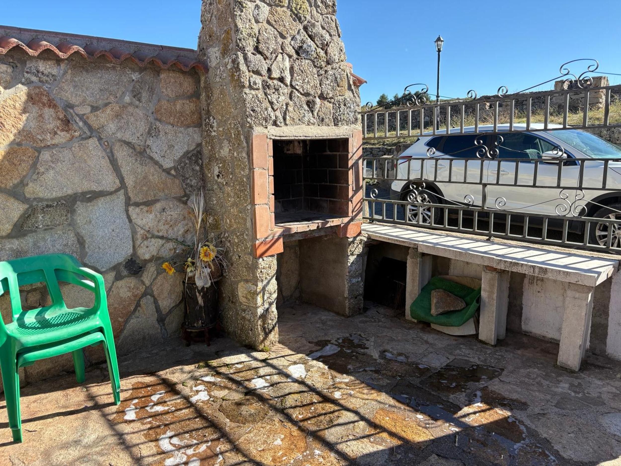 A stone wall with a small opening, a green plastic chair, and a car parked behind a metal fence.
