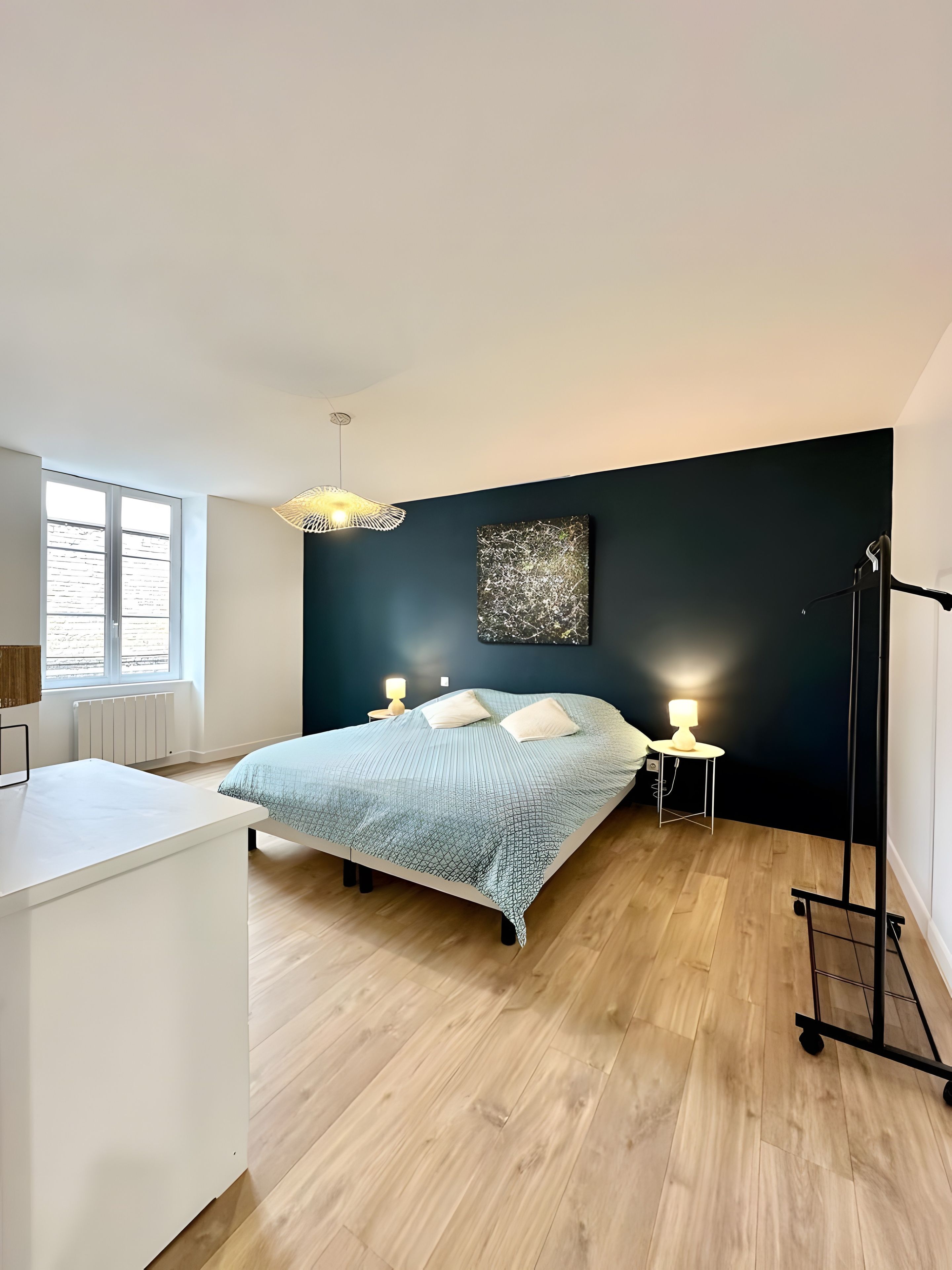 Ein modernes Schlafzimmer mit einem Bett, Nachttischlampen, einem Heizkörper, einem Fenster und einer schwarzen Wand mit strukturierter Dekoration.