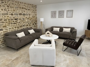 A living room with a stone wall, two sofas, a coffee table, a white armchair, and a TV stand.