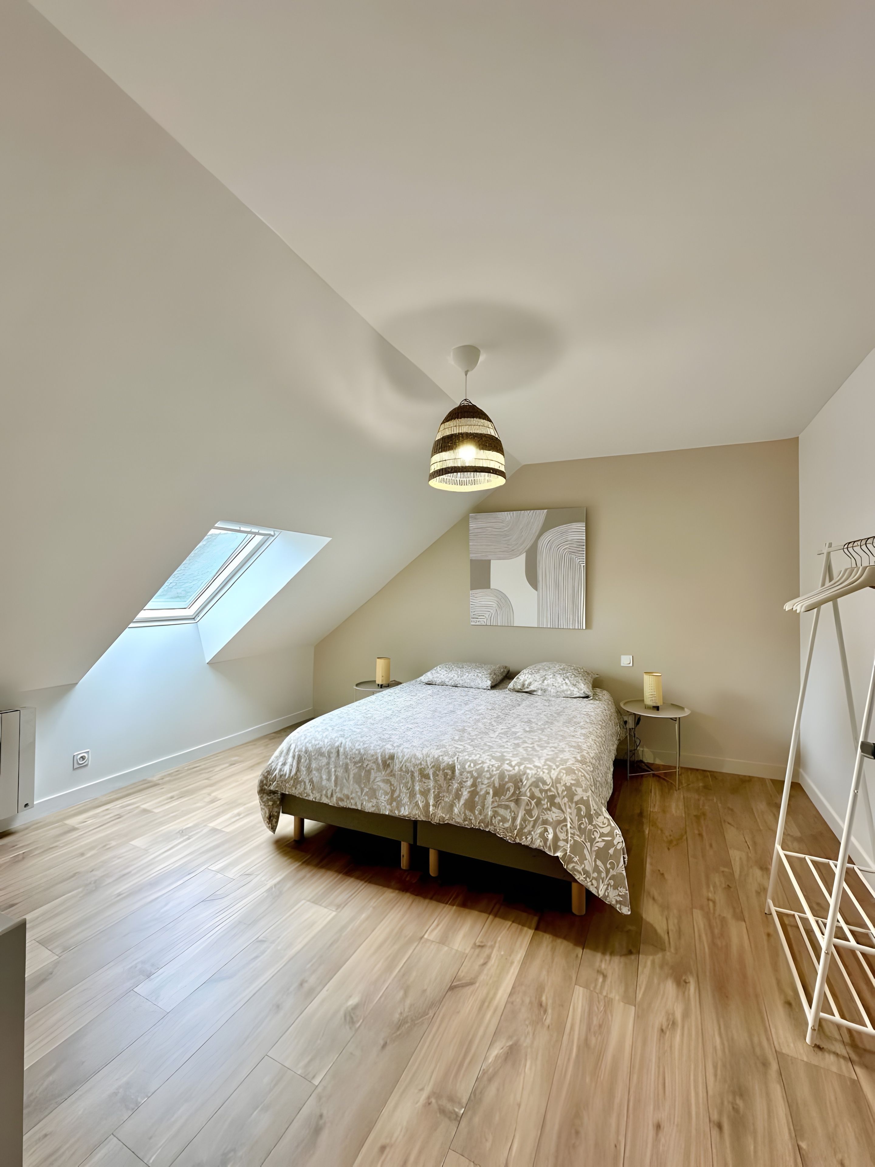 Une chambre avec un plafond en pente, un lit recouvert d’une couvre-lit à motifs, un luminaire suspendu, une petite table de chevet avec une lampe et un puits de lumière.