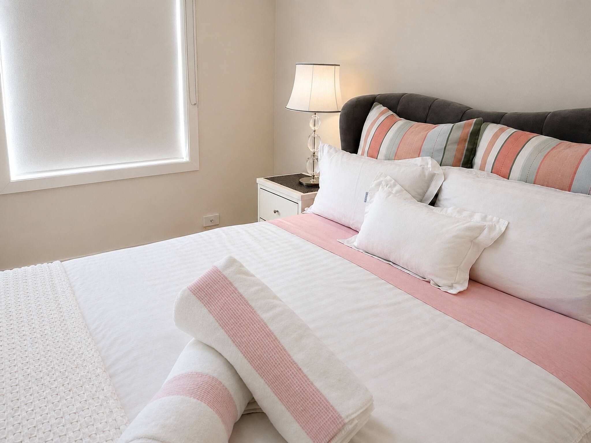 Un lit bien fait, avec une literie blanche et rose, une lampe de chevet et un coussin à rayures.