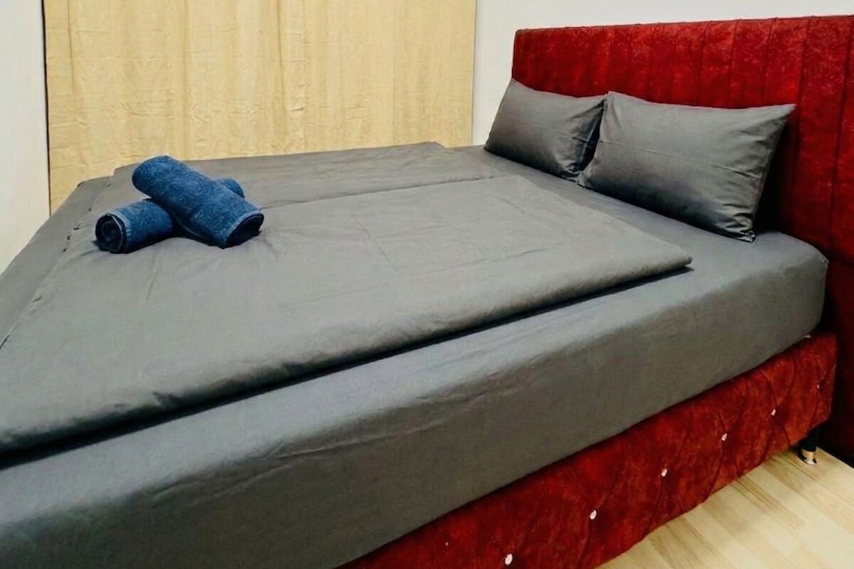 Una cama individual con colchón gris y cabecera roja, un par de toallas azules enrolladas y cortinas color beige en una pared blanca.