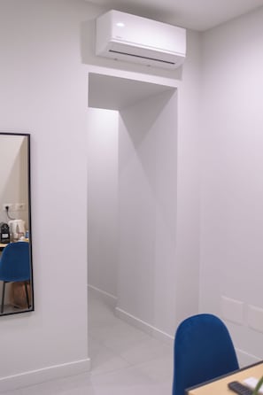 A room with a white air conditioning unit mounted on the wall, a blue chair, a table with a kettle, and a mirror reflecting part of the room. - The Ease (Naples)